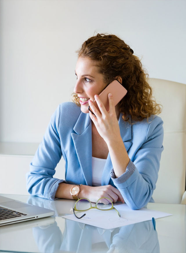 CBS Konsult Person in a blue blazer sitting at a desk, holding a phone to the ear, with a laptop, glasses, and papers on the desk.